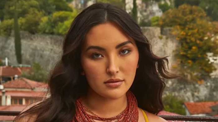 Yumi Nu poses in front of a natural stone backdrop wearing a red beaded necklace and her dark brown hair down in a soft wave.